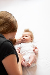 Caring young mother playing with her newborn baby daughter in the bed and looking at her with smile, happy moments of maternity
