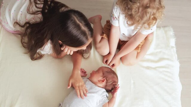 Joyful sisters wearing T-shirts carefully cuddling their little brother whispering softly to him entertain their baby brother in cozy bedroom