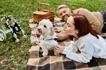 A man and woman are laying on a blanket with their dog in a peaceful park setting.
