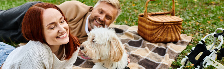 A man and woman relax on a cozy blanket with their dog in a park.