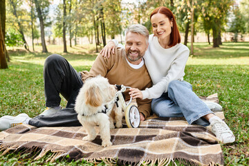A man and woman relax on a blanket with a dog in a peaceful park setting.