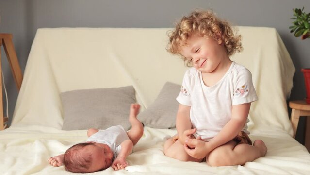 Childcare and sibling love. Smiling blond wavy haired older sister playing with her newborn baby brother sitting on white sofa in living room loving sister with baby