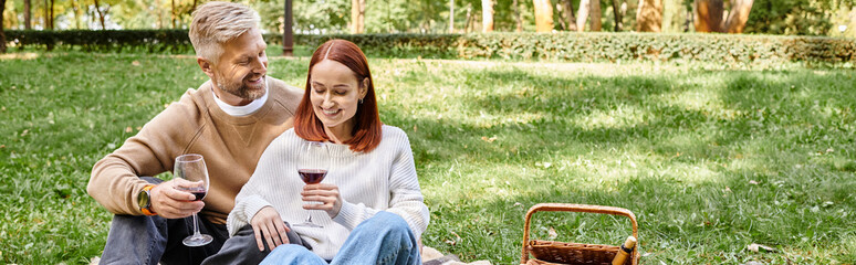 Adult man and woman sitting on a blanket, holding wine glasses in a park.