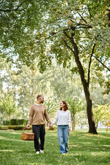 Obraz premium Man and woman in casual attire holding hands, walking through park.