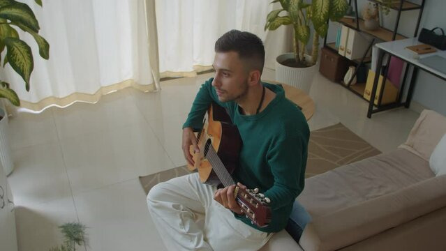 Medium long shot of Caucasian male songwriter creating melody with acoustic guitar at home