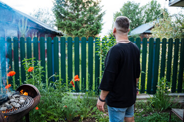 A man is grilling at a backyard BBQ surrounded by blooming flowers and a fence, enjoying the summer outdoors with friends and family in a relaxed and casual setting