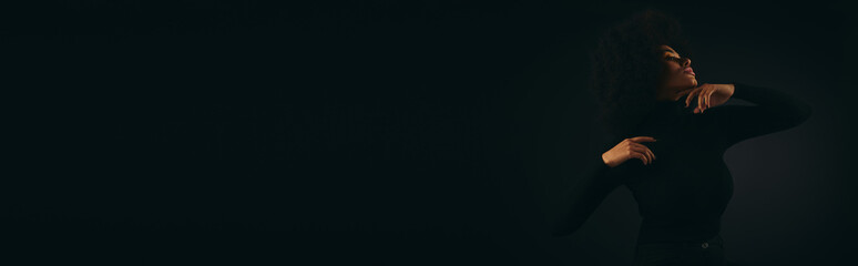 Stylish African American woman stands with arms outstretched in darkness.