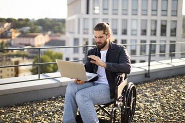 Fototapeta premium Caucasian man in wheelchair using laptop on city rooftop. Concept of remote work, determination, and modern lifestyle.