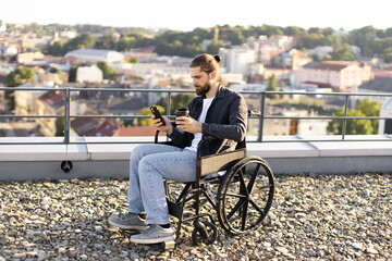 Young man in wheelchair using smartphone on rooftop overlooking cityscape. Concept of accessibility, modern technology, and urban lifestyle.