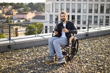 Fototapeta premium Man on rooftop in wheelchair using smartphone and holding coffee cup. Urban background with buildings and greenery.