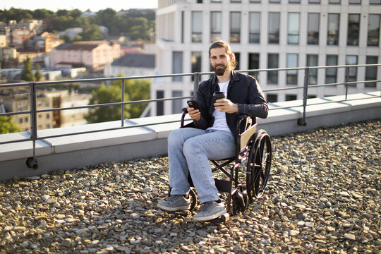 Man on rooftop in wheelchair using smartphone and holding coffee cup. Urban background with buildings and greenery.