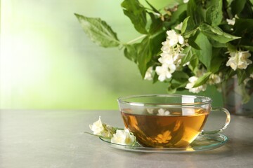 Aromatic jasmine tea in cup, flowers and green leaves on light grey table, space for text