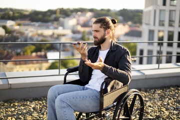 Young man in wheelchair talking on smartphone while on rooftop. Urban cityscape background. Concept of communication and accessibility.
