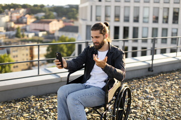 Obraz premium Young man in wheelchair making video call on rooftop terrace, engaging in an online conversation, showcasing modern technology and accessibility.