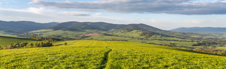 A winding path cuts through a lush green field to the Kralicky Sneznik Mountains of Czechia, offering a scenic view of the rolling hills under a blue sky.