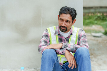 Asian male architect in reflective vest and hard hat sitting on concrete floor. Looking tired,...