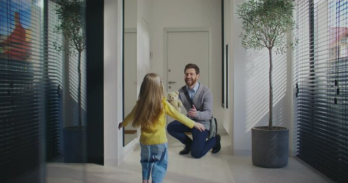Young girl running fast towards her Caucasian father. Man came home after work or long business trip. Holding toy for his beloved child. Hugging each other. Sun shining bright through window.