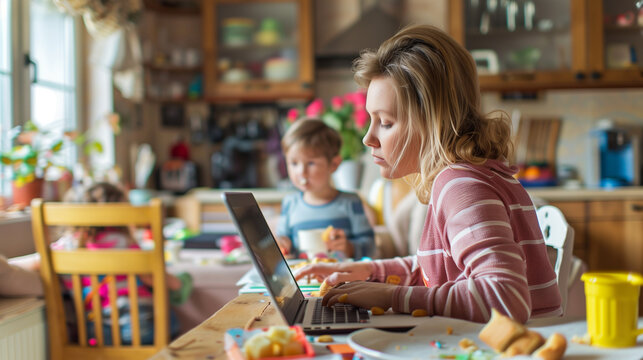 Young mother is multitasking, working from home on her laptop while her children are having breakfast in the kitchen - Powered by Adobe