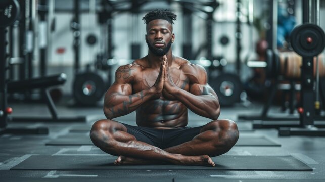A muscular man sits in a meditative pose on a mat in a modern gym, surrounded by weights and exercise equipment.