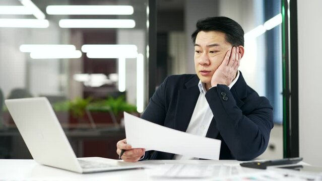 Overworked asian businessman is tired doing paperwork and working on a laptop computer sitting at workplace in business office. A mature man in a formal suit is overtired and bored. Overload at work