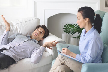 A man lies on a white sofa, animatedly sharing his thoughts with a female therapist who is attentively taking notes. The setting is a cozy living room, featuring a bright, calm ambiance.