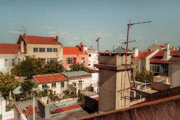 Lisbon cityscape featuring traditional residential buildings with red tiled roofs, visible antennas, and lush gardens. A clear blue sky and sunlight highlight the urban neighborhood.