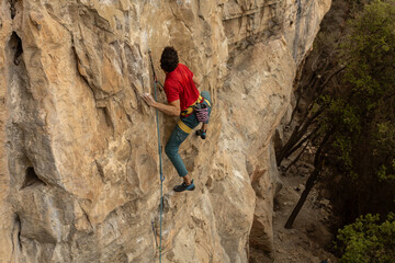 A man is climbing a rock wall with a blue rope. The man is wearing a red shirt and yellow pants