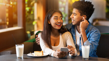 A young black couple enjoys sharing earphones and listening to music while sitting at a cafe table, smiling and laughing together on a sunny day. Each has a glass of milk and a slice of cake