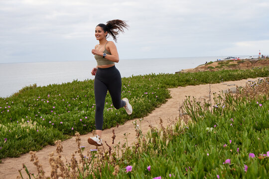 Hispanic female runner running along a dirt path with the ocean in the background, wearing dark tights and olive top.