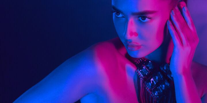 A woman is bathed in blue and pink neon lighting at night. She is touching her face with one hand while gazing intently into the distance, creating a dramatic and captivating visual effect.