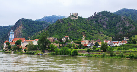Fototapeta premium Panoramic Scenic view while cruising along the River Danube in Austria. Pretty waterfront village with blue church and quaint cottages, all nestled amongst lush green mountains.