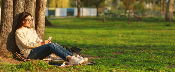 Asian young woman enjoys a peaceful afternoon in the park, sitting against a tree, and checking her...