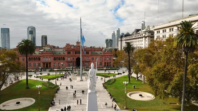 Dozens of people walk through Plaza de Mayo. Aerial camera moves forward towards Casa Rosada flying over the central monument