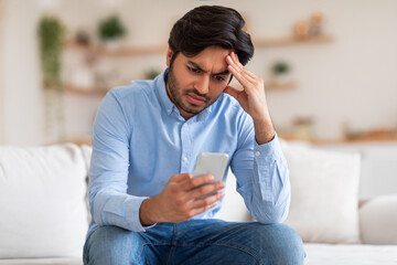 A young man with a concerned expression sits on a couch while checking his smartphone. He is...