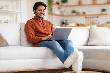 Obraz premium A man in a casual orange shirt and blue jeans sits comfortably on a white couch with his legs extended and a laptop in his lap. He is smiling as he types, seemingly working on a project