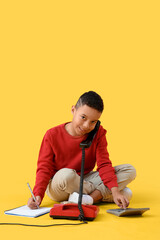 Little African-American boy talking by telephone while writing in notebook on yellow background