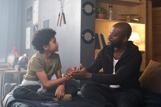 Side view portrait of caring African American father talking to son and holding hands sitting on bed together