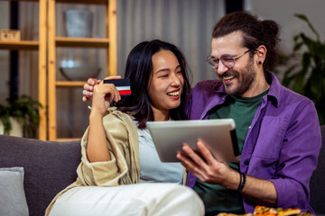 A couple, an Asian woman and a white man, is comfortably seated in their cozy living room. They are engaged in making an online payment together,