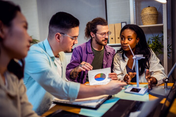A group of four young people from diverse backgrounds gather around a model wind turbine. They share their thoughts and ideas about sustainable energy sources. 