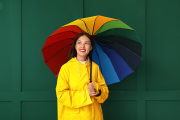 Beautiful young Asian woman in raincoat with umbrella on green background