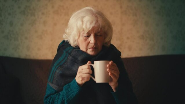 Poor senior woman wrapped in blanket trembles from cold at home, drinks hot tea