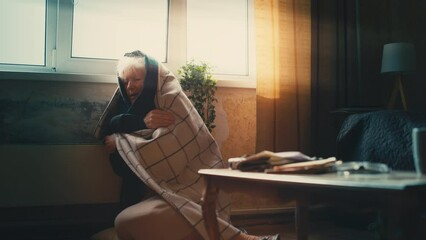 Upset aged woman wrapped in blanket warming up near electric heater, cold room