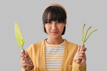 Beautiful young happy female gardener with gardening rake and shovel on grey background