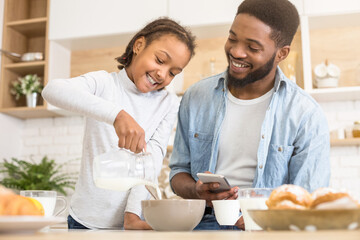 A young African American girl is pouring milk into a bowl while her father, holding a cellphone, watches with a smile in a kitchen setting