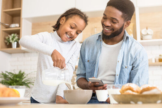 A young African American girl is pouring milk into a bowl while her father, holding a cellphone, watches with a smile in a kitchen setting