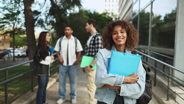 Portrait of a smiling young latin student looking at the camera while her classmates talk in the background on campus. Students socializing outdoors on campus, one smiling with folders