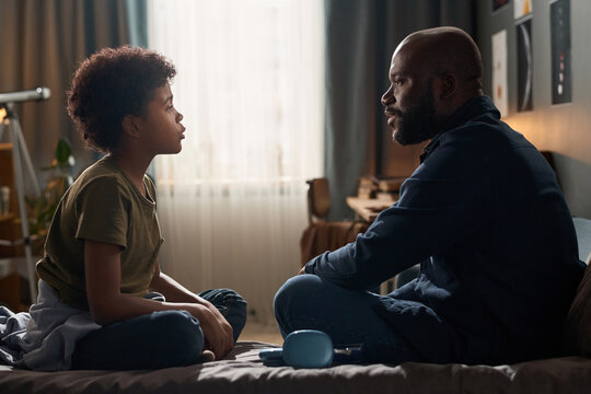 Side view portrait of African American family with single father and son talking to each other sitting on bed copy space