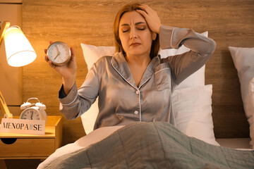 Mature woman with alarm clock cannot sleep in bedroom at night. Menopause concept