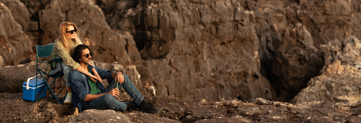 A couple enjoys the sunset on a rocky outcrop. The woman is seated in a folding chair, while the man sits on the rocks. They appear to be relaxing and enjoying the view, copy space