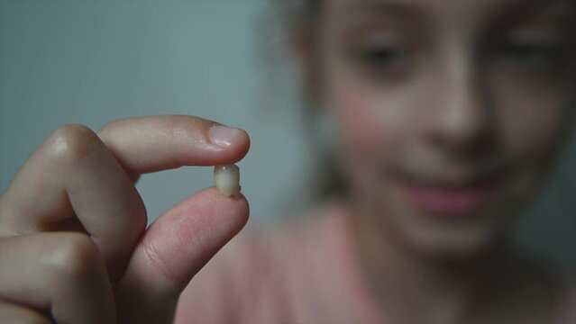 A little girl, 8-9 years old, has lost a baby tooth and she holds it with her fingers, showing to the camera